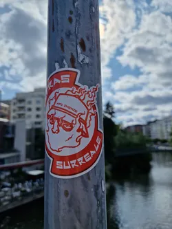 Red and white sticker featuring a surreal illustration of a face with flames, and the word 'Surreale' at the bottom. The text 'HPC CHEMISE' is visible on the top of the illustration. The sticker is attached to a gray metal pole outdoors, with a body of water visible in the background.