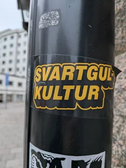 The sticker is predominantly black with bold, yellow lettering. The text is arranged in two lines: 'SVARTGUL' on the top line and 'KULTUR' on the bottom line. The letters have a thick, black outline, and the yellow fill has a slightly textured appearance. The sticker has a wavy, irregular edge, suggesting it's a die-cut sticker. It appears to be adhered to a black cylindrical pole.