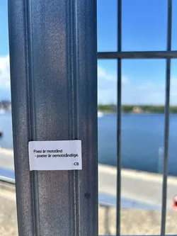 Street sticker A small, rectangular white sticker featuring black sans-serif text in Swedish. The sticker is affixed to a grey metal fence post with a blurred waterfront background.