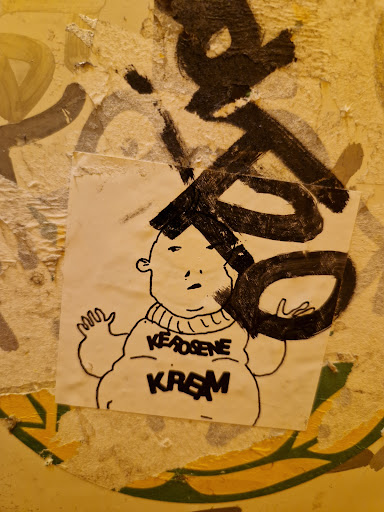 Street sticker A sticker featuring a cartoon drawing of a chubby character wearing a sweater with the words "Kerosene Kream" written on it. The drawing is in black ink on white paper. The sticker is attached to a wall with other graffiti and markings.