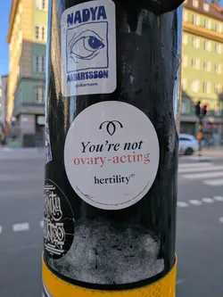 Street sticker A white circular sticker with black and red text reading 'You're not ovary-acting'. It features a minimalist ovary icon at the top and the brand name 'hertility' at the bottom, along with a website URL in fine print.