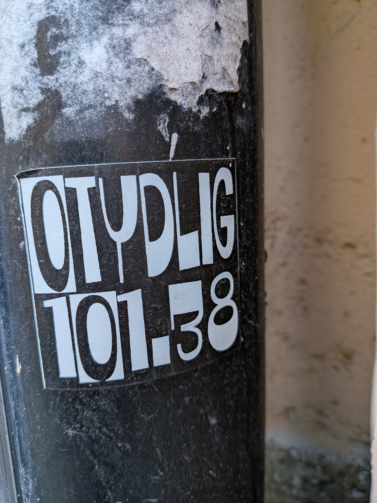 A rectangular sticker featuring a stark black background with prominent white (or very light grey) text. The top line displays the word 'OTYDLIG' in a bold, sans-serif, slightly rounded block font. Below it, the numbers '101.38' are presented in a similar, though somewhat more condensed, bold typeface. The sticker is applied to a dark, textured pole and exhibits signs of wear, including dirt accumulation and slight peeling along its upper edges.