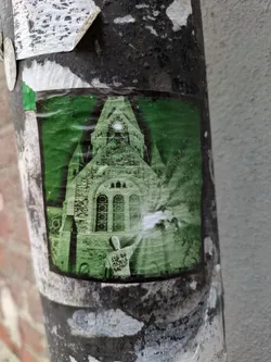 This sticker features a monochrome image of a person seemingly radiating light in front of a church. The image is in shades of green and has a slightly faded quality, adding to its mysterious atmosphere. The text on the figure's back is partially visible but illegible. The sticker shows signs of wear, indicating it has been exposed to the elements for some time.