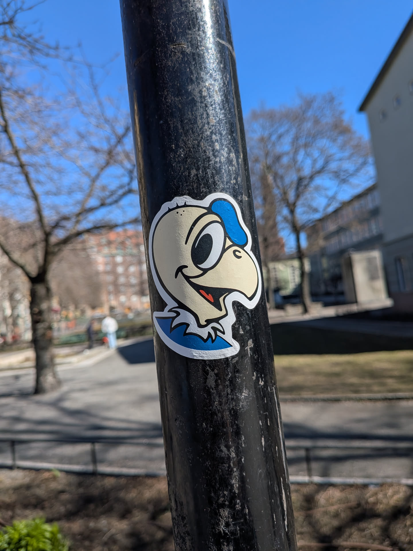 The sticker features a cartoon character's head and upper neck, resembling a bird or a creature with a large beak. The character has a light beige or cream-colored head with large, round white eyes and black pupils, giving it a wide-eyed, friendly expression. A prominent, slightly upturned beak is visible. The mouth is open in a wide smile, revealing a red tongue and a black interior. A blue, somewhat amorphous shape sits on top of the head, possibly a cap or stylized hair, and a matching blue, ruffled collar-like element is around the neck. All elements are outlined with thick black lines, and there are a few small black dots on the head. The entire character is set against a white border, indicating it's a die-cut sticker applied to a textured black metal pole.