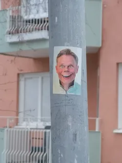 Street sticker The sticker is a rectangular, vertically oriented image featuring a man's headshot. He has light brown hair, fair skin, and a very wide, almost exaggerated, toothy smile. His blue eyes are wide open, contributing to an intense or slightly unsettling expression. He is wearing a light-colored, possibly mint green or light blue, collared shirt with a zipper at the neck. The background of the portrait is plain white. The sticker is affixed to a grey, textured metal pole.