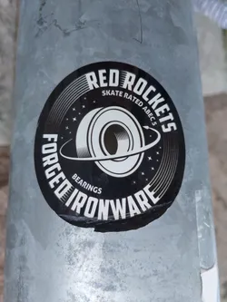 Street sticker A black and white circular sticker with the text "Red Rockets Forged Ironware" written around a graphic of a bearing. The text "Skate Rated ABEC 5" is written in smaller font above the bearing graphic. The sticker has a worn appearance.