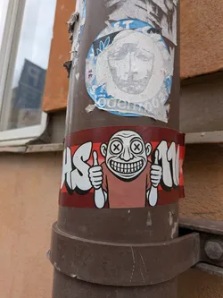 A rectangular sticker wrapped around a brown pipe, featuring a central cartoon character with a bald head, large ears, 'X' eyes, and a wide, toothy grin. The character is wearing a reddish-brown shirt and giving a double thumbs-up. The background has horizontal red stripes, with white bubble letters 'AS' on the left and '110' on the right, both outlined in black.