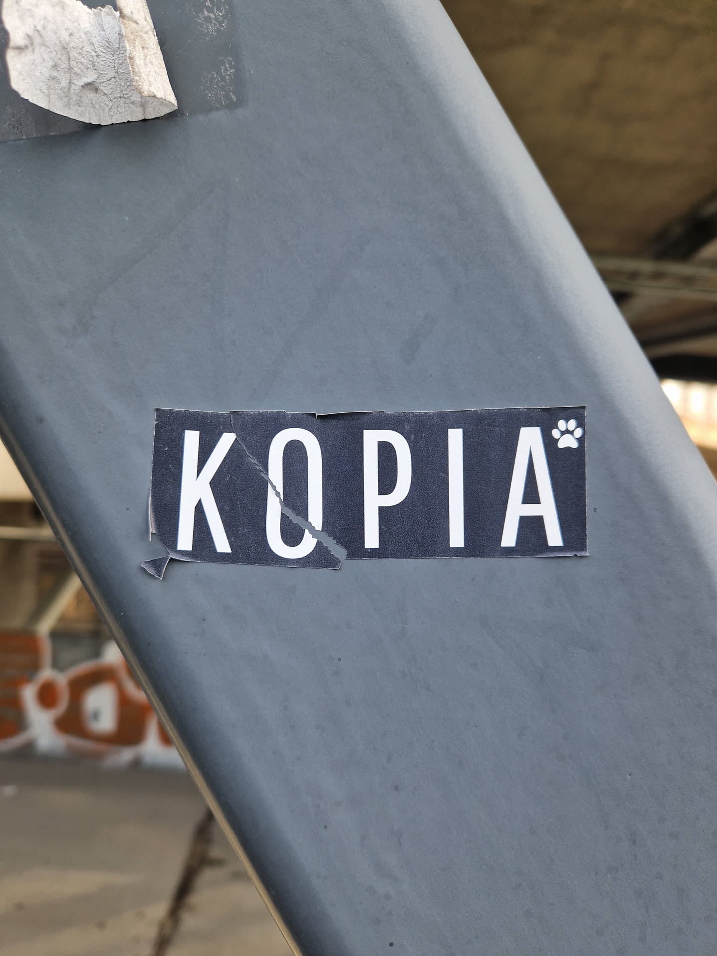 A rectangular sticker with a black background and bold, white, sans-serif text that reads 'KOPIA'. A small white paw print icon is positioned at the top right of the letter 'A'. The sticker shows signs of wear, including a diagonal tear through the letter 'O' and peeling edges on the left side. It is stuck onto a smooth, dark grey metal surface.