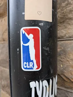 Street sticker The sticker is a rounded rectangular shape, mimicking the iconic NBA logo. It features a white silhouette of a person holding a spray can, reaching upwards, set against a split background of vibrant blue on the left and bright red on the right. The person's pose is a clear homage to the NBA's Jerry West silhouette, but recontextualized with a graffiti artist's tool. At the bottom left, within the blue section, the white capital letters 'CLR' are prominently displayed. The sticker has a thin white border around its perimeter.