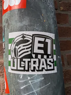 A rectangular sticker with a white border, featuring a background of alternating horizontal green and white stripes. On the left, a stylized black and white emblem resembling a laurel wreath or crest with horizontal stripes. To its right, the bold, blocky letters 'E1' are outlined in black and filled with black. Below these elements, the word 'ULTRAS' is prominently displayed in large, blocky white letters with a thick black outline. The overall design is stark and graphic, primarily in black, white, and green.