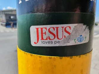A sticker on a pole with the text "JESUS loves pe..." and a menorah symbol. The sticker is partially torn.