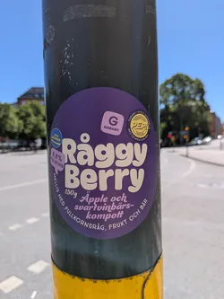 Street sticker The sticker is circular with a vibrant dark purple background. The main text, 'Råggy Berry', is prominently displayed in a large, playful, light cream-colored font across the center. Below it, in a smaller, italicized cream font, is '150g Äpple och svartvinbärs-kompott'. Along the bottom edge, also in cream text, it reads 'MELLIS MED FULLKORNSRÅG, FRUKT OCH BÄR'. On the left side, a light purple speech bubble contains '8.8% FULLKORN' in cream text, and above it, a blue and yellow circular logo states 'FRÅN SVERIGE'. On the top right, there's a square-ish light purple logo with a cream 'G' and 'GARANT' below it. To its right, another circular logo in yellow and black reads 'GENERATION PEP SWEDEN' and 'GARANT I STOLT SAMARBETE MED GENERATION PEP'. The sticker is affixed to a dark grey pole.