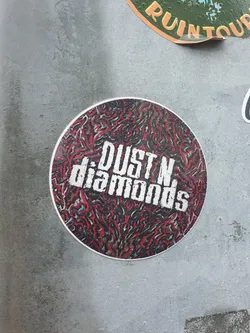 Street sticker Round sticker with a red, black and green abstract pattern. The text Dust N Diamonds is written in white uppercase letters in the center.