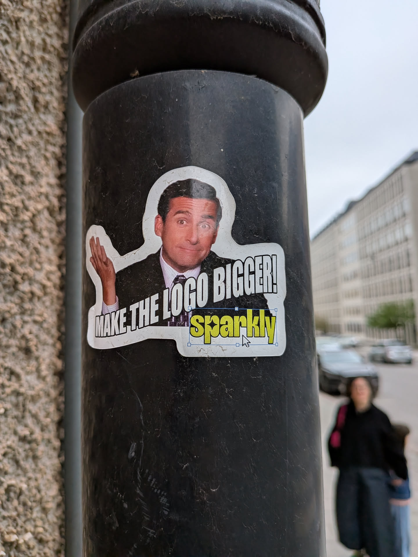 A die-cut sticker featuring a cutout image of Michael Scott (Steve Carell) from 'The Office' in a suit and tie, with his right hand raised in a gesturing manner. He has a slightly bewildered or emphatic expression. Below his image, in large white block letters with a black outline, is the text 'MAKE THE LOGO BIGGER!'. Directly beneath this, the word 'sparkly' is written in a vibrant yellow-green color with a black outline, appearing as if it's being edited in a graphic design program, complete with a blue selection box and a white mouse cursor icon pointing to it. The sticker has a clean white border around the entire image and text, and it is affixed to a dark, textured pole.