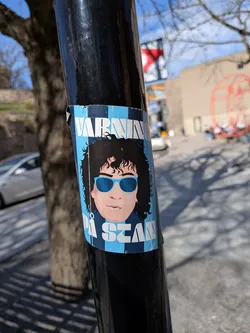 Street sticker A rectangular sticker adhered to a black pole, featuring a stylized cartoon-like depiction of a person from the shoulders up. The character has dark, voluminous curly hair and is wearing bright blue sunglasses. Their skin tone is a light peach. The background of the sticker is composed of alternating light blue and dark blue vertical stripes, with a checkerboard pattern visible at the very top. White block letters are overlaid on this striped background. The sticker shows signs of wear, with slight peeling and creasing, particularly at the top and bottom edges, indicating it's a physical sticker exposed to the elements.