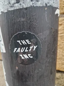 A round black and white sticker with the words "THE FAULTY INC" in a stylized font. The sticker is affixed to a dark gray or black textured surface, possibly a pole or pipe.