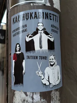 Street sticker A sticker featuring a stylized illustration of three individuals.  A man in a Dracula-like cape is centrally positioned, flanked by a woman on the left and another woman on the right.  Text in Finnish surrounds the figures, suggesting a theme of horror or satire related to wellbeing, poverty, and art.