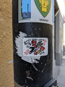 Street sticker A rectangular sticker with rounded corners featuring a vibrant, cartoonish illustration on a white background. The main figures are a red, horned, devil-like character with a black body, white gloves, and red shoes, holding what appears to be a tattoo gun. Beside it is a black animal, possibly a badger or a similar creature, with white facial markings, holding a green object. Both characters have dynamic poses with red lines radiating from them, suggesting motion or energy. Above the characters, the text "LOCAL. MAN. TATO" is written in green and black. Below the characters, the word "TATTOO" is prominently displayed in large, colorful letters (red, yellow, green, blue), followed by three black asterisks. The overall style is bold, playful, and edgy.
