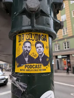 Street sticker A rectangular yellow sticker affixed to a dark green metal post. The sticker features black-and-white portraits of two men, Matias Varela on the left and Calle Schulman on the right. The background is yellow and decorated with various black line doodles, including stars, a 'Cool S', and crude drawings. The text 'DE OLYDIGA' is written in a stylized black font at the top, and 'PODCAST' along with 'MATIAS VARELA & CALLE SCHULMAN' is printed at the bottom.