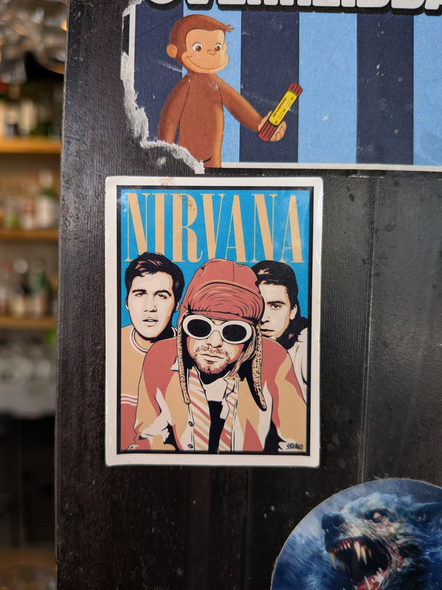 A rectangular sticker featuring a stylized illustration of the band members of Nirvana. Kurt Cobain is centered in his signature white sunglasses and red hat, with Krist Novoselic on the left and Dave Grohl on the right. The band name 'NIRVANA' is written in large yellow letters at the top against a blue background.