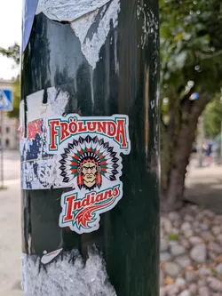 Street sticker Die-cut sticker of the Frölunda Indians logo on a dark pole. Features a Native American chief in a feathered headdress, with 'FRÖLUNDA' arched above and 'Indians' below in red and green.