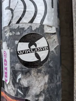 The sticker is circular, featuring the iconic Wu-Tang Clan 'W' logo in white, set against a black background. Horizontally across the center of the 'W' logo, the word "WIKLUND" is written in white, in a stylized, somewhat gothic or old English-style font. The sticker appears to be slightly worn and dirty, consistent with being affixed to an outdoor pole, showing some scuffs and minor peeling.