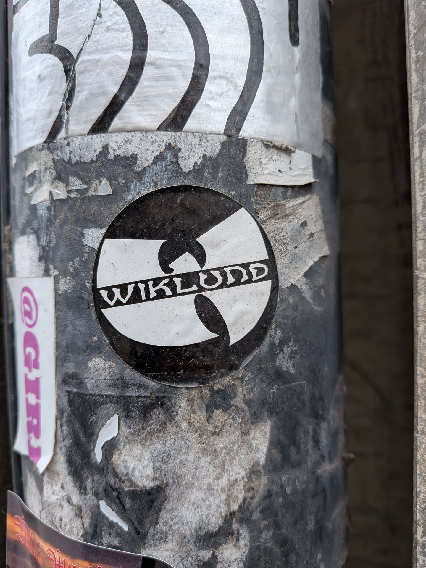 The sticker is circular, featuring the iconic Wu-Tang Clan 'W' logo in white, set against a black background. Horizontally across the center of the 'W' logo, the word "WIKLUND" is written in white, in a stylized, somewhat gothic or old English-style font. The sticker appears to be slightly worn and dirty, consistent with being affixed to an outdoor pole, showing some scuffs and minor peeling.