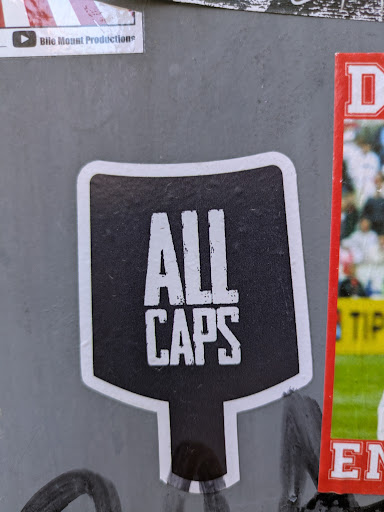 The sticker is black and white. It has a rounded square shape on top, with a stem coming out from the bottom. The text on the sticker says "ALL CAPS".