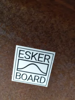 A white square sticker with black text and a graphic of a hill. The text on the sticker says "ESKER" above a graphic of a hill inside a box and "BOARD" below the graphic.