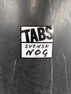 Street sticker The sticker is rectangular with a white background and black text. The top section prominently features the word 'TABS' in large, bold, sans-serif letters. Below 'TABS', in a slightly smaller font, is the word 'SVENSK', followed by 'NOG' in a large, distinct, handwritten or brush stroke style font. At the very bottom, in small, uppercase sans-serif text, it reads 'STREAMA NU'. The sticker itself appears slightly worn around the edges and is affixed to a dark, possibly metallic or plastic, textured surface with some white scuff marks.