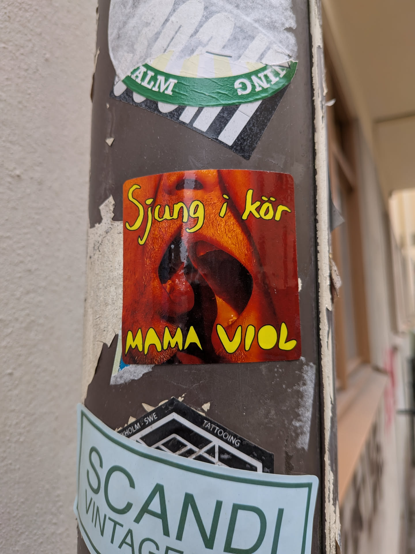 A rectangular sticker with rounded corners depicting a close-up, warm-toned photograph of a wide-open mouth, as if singing or shouting. The image has a strong orange and red cast. At the top, the Swedish phrase 'Sjung i kör' (Sing in a choir) is written in a stylized yellow font with a black outline. At the bottom, the words 'MAMA VIOL' are written in the same yellow font.
