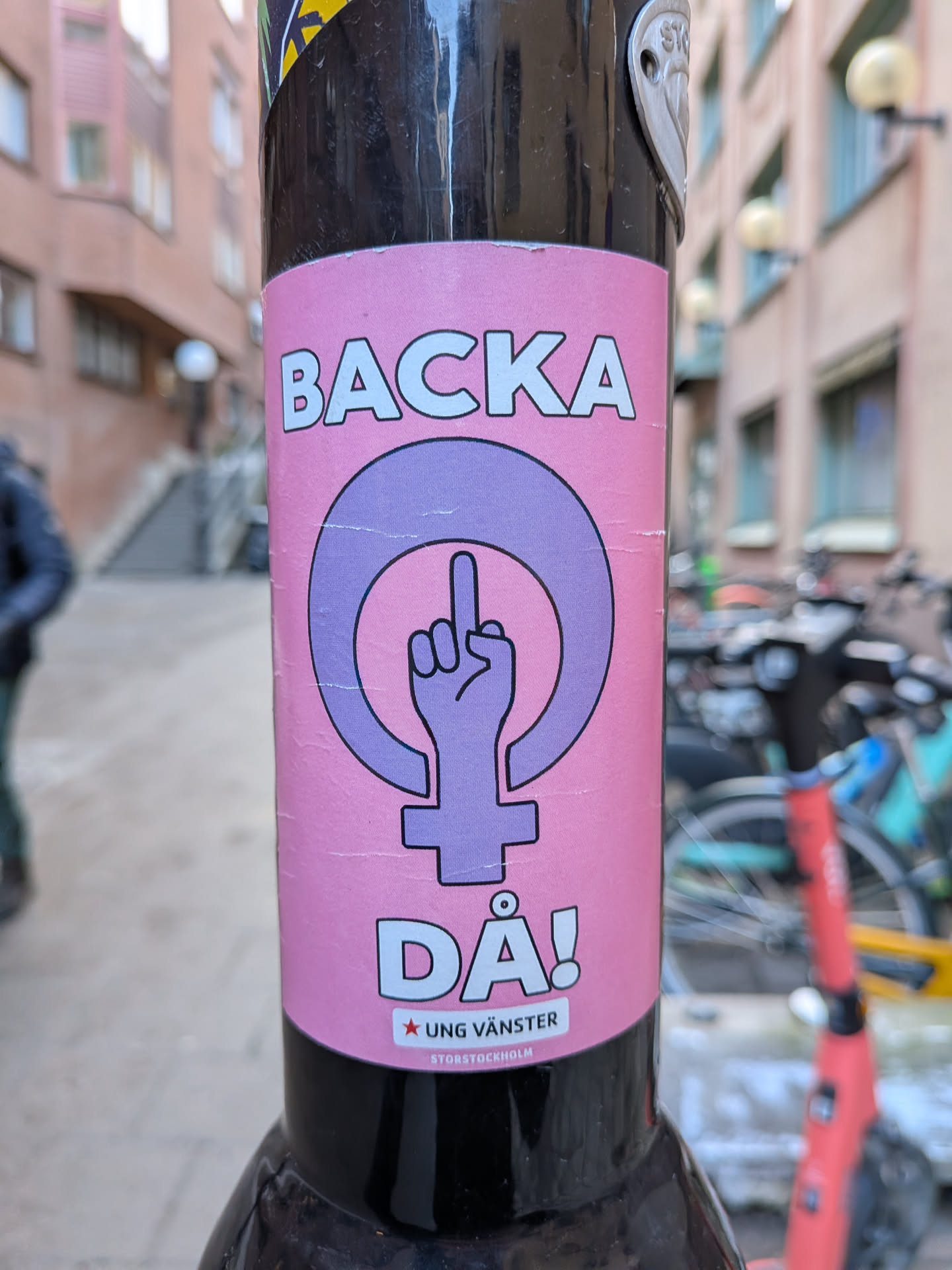 A rectangular pink sticker adhered to a black pole. The sticker features a large purple Venus symbol (female symbol) centrally placed. Inside the circle of the Venus symbol, a hand is depicted giving the middle finger, outlined in black and filled with a lighter shade of purple. Above the symbol, the word 'BACKA' is written in large, bold white letters with a black outline. Below the symbol, 'DÅ!' is written in the same style. Further below, a small red star precedes the text 'UNG VÄNSTER' in white letters on a light teal background strip. At the very bottom, in smaller white text, is '@STORSTOCKHOLM'. The sticker shows minor creases and wear, indicating it's a physical object.