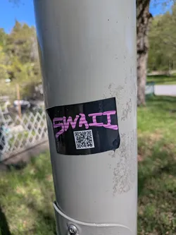 A rectangular sticker with a black background adhered to a light grey pole. The sticker features the word 'SWAII' in a bold, stylized, somewhat graffiti-like pink font across the top. Below the text, a white QR code is prominently displayed against the black background. The sticker shows minor signs of wear, with slightly curled edges, particularly on the top left and bottom right.