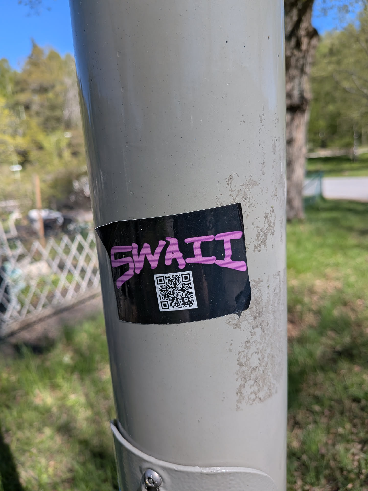 A rectangular sticker with a black background adhered to a light grey pole. The sticker features the word 'SWAII' in a bold, stylized, somewhat graffiti-like pink font across the top. Below the text, a white QR code is prominently displayed against the black background. The sticker shows minor signs of wear, with slightly curled edges, particularly on the top left and bottom right.