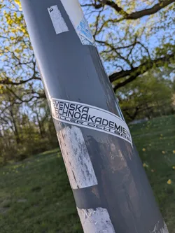 Street sticker The sticker is a horizontal rectangular design with a clean white background and bold black sans-serif text. It features two lines of text: 'SVENSKA TECHNOAKADEMIEN' is prominently displayed on the top line, with 'RULLER OCH BAS' on the line below. The sticker is affixed to a dark grey, cylindrical pole, showing minor signs of wear and tear, including slight peeling at the edges and some dirt accumulation, indicating it is a genuine, weathered sticker.