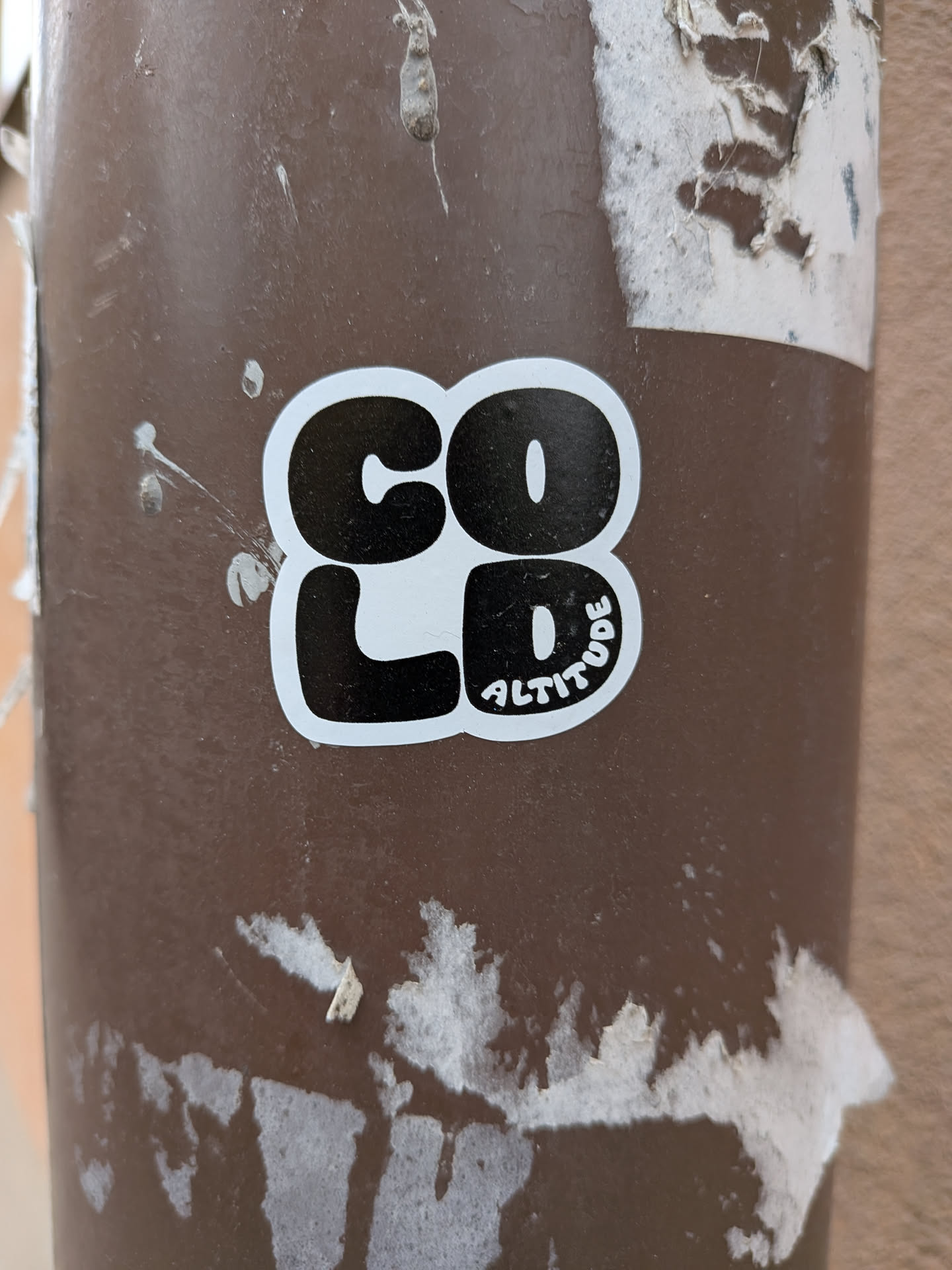 A die-cut sticker with a white border featuring the word 'COLD' in large, bold, black bubble letters arranged in a two-by-two grid. The letters 'C' and 'O' form the top row, while 'L' and 'D' form the bottom row. Inside the bottom curve of the letter 'D', the word 'ALTITUDE' is written in small, white, sans-serif capital letters. The sticker is affixed to a weathered brown metal pole.