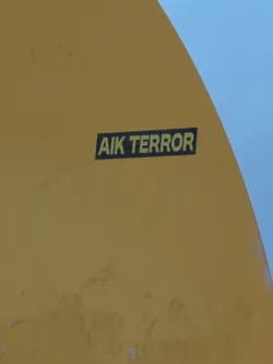 A rectangular sticker featuring the text 'AIK TERROR' in a bold, sans-serif, yellow-green font against a solid black background. The sticker is applied to a curved, mustard-yellow surface, possibly part of a vehicle or structure, with a clear blue sky visible in the upper right corner. The design is simple, high-contrast, and direct.