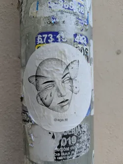 This round sticker is adhered to a textured, weathered pole covered in layers of other stickers and peeling posters. The sticker's design is a stylized black and white drawing of a face with butterfly wings in place of some features. The face appears to have one tear, and there is text at the bottom of the sticker.