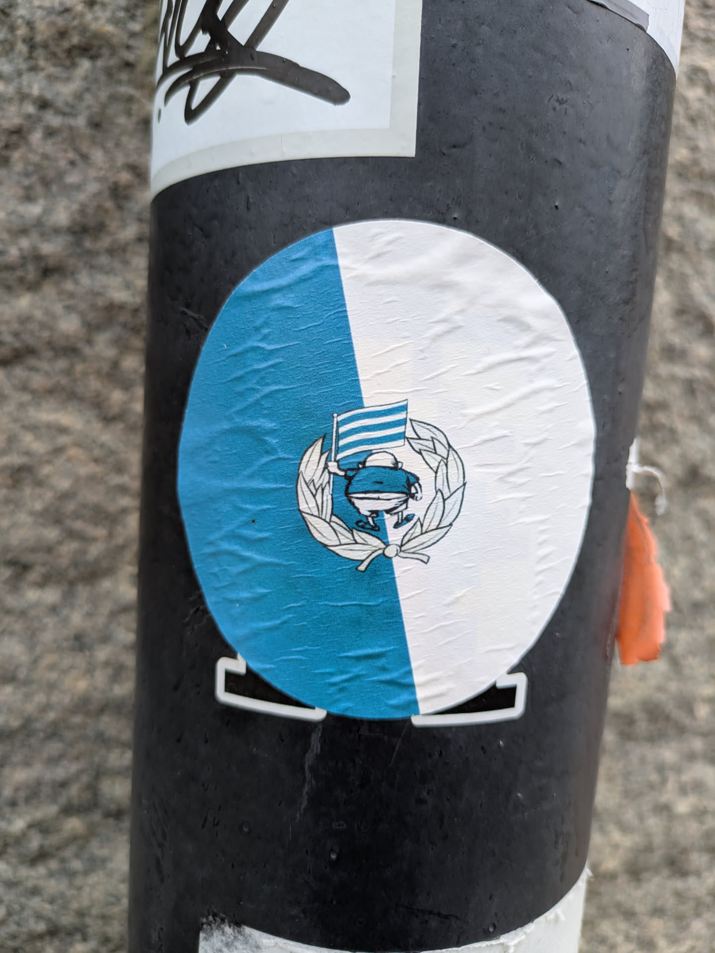 The sticker, affixed to a black pole, features a round emblem split vertically into blue and white halves. In the center of the emblem is a cartoon-style frog figure holding a flag. The frog and flag are encircled by a laurel wreath. The flag has horizontal lines and is also stylized.