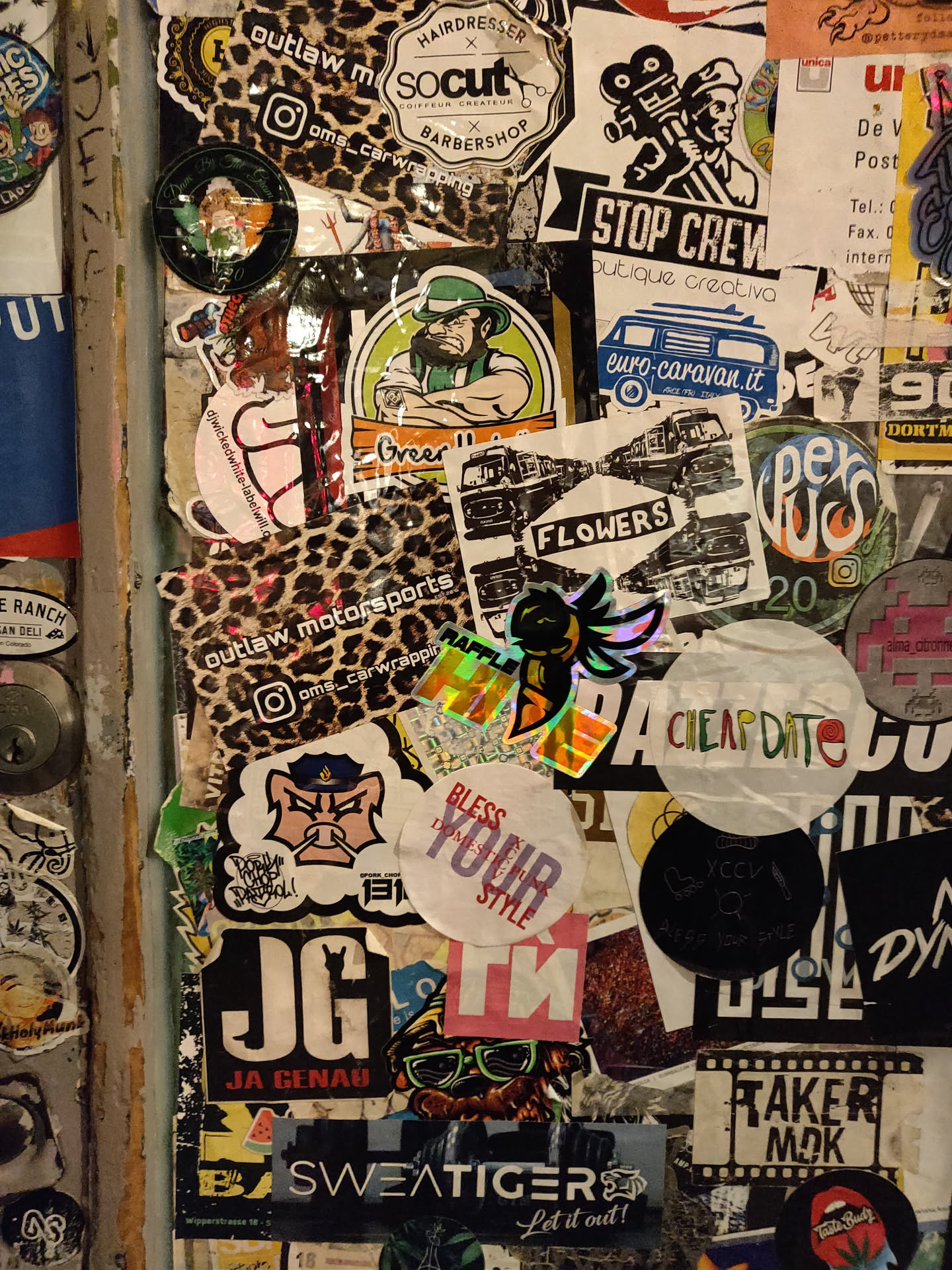 Close-up view of a cluster of stickers, predominantly on a dark-colored surface.  The stickers vary in shape, size, design, and color.  One prominent sticker displays a holographic or iridescent effect. Some stickers feature text, logos, and illustrations.