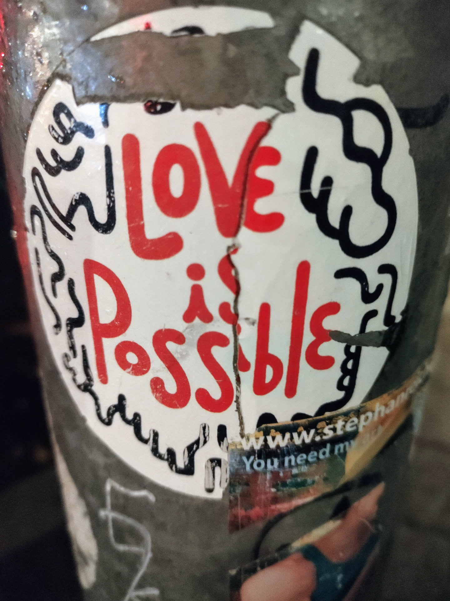 A sticker with the text "Love is Possible" written in red on a white background. The sticker is slightly damaged and has some black doodles around it. There's a partially visible smaller sticker underneath with a website address and text.
