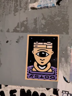 The sticker is rectangular with a light orange-beige border, framing a central image on a black background. The main subject is a stylized, light beige head featuring a prominent single eye with a large black pupil and a small white highlight, and a simple, curved mouth below it. The head wears a distinctive, multi-layered headpiece in beige and white. The figure appears to be emerging from or submerged in a purple, wavy, or patterned surface at the bottom. Small purple dots are scattered across the black background above the head. The word "DOTSY" is written in white, inverted, across the bottom of the purple section. The overall aesthetic is clean, graphic, and illustrative.