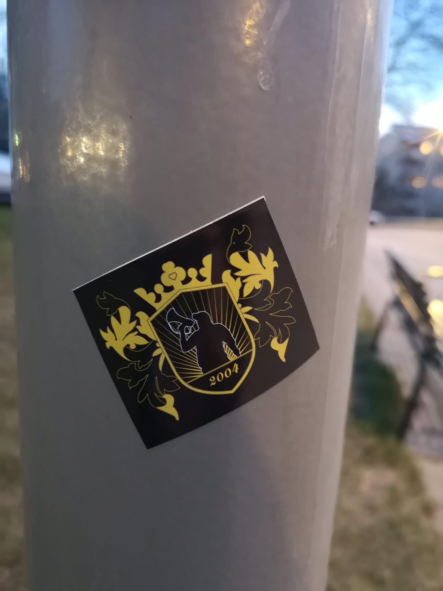 The sticker is square-shaped with a black background and features intricate yellow/gold designs. At its center is a shield or crest, within which a black silhouette of a person holding a megaphone is depicted, facing right. Behind the figure, yellow/gold rays emanate. Below the silhouette, the year '2004' is written in yellow/gold. The shield is topped with a stylized crown, which includes a small heart symbol at its apex. Elaborate yellow/gold floral or leafy scrollwork flanks the central crest, giving the entire design a regal or heraldic appearance. The sticker is applied to a light grey pole.