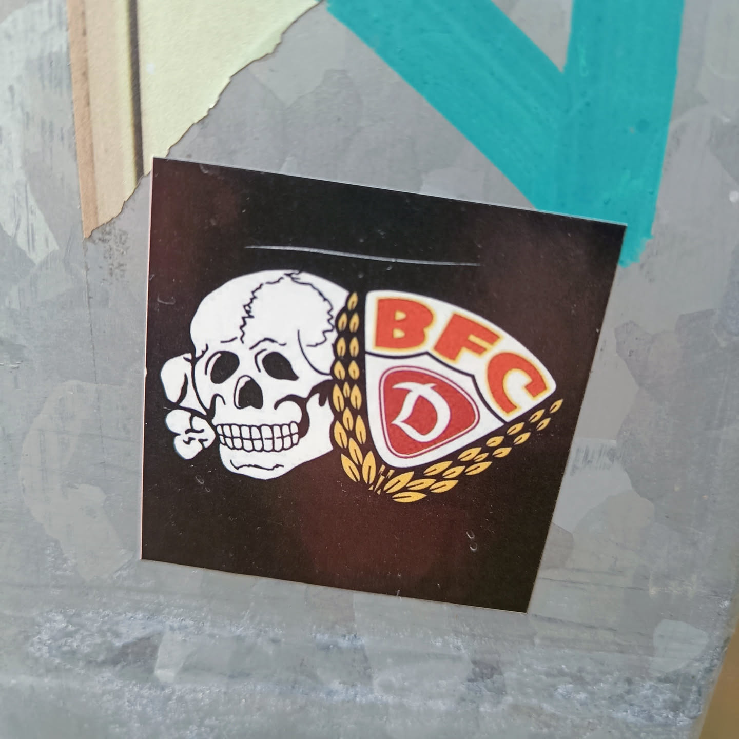 A black sticker with a skull and the logo of BFC Dynamo, a German football club. The skull is depicted in white and the logo is in red and gold.