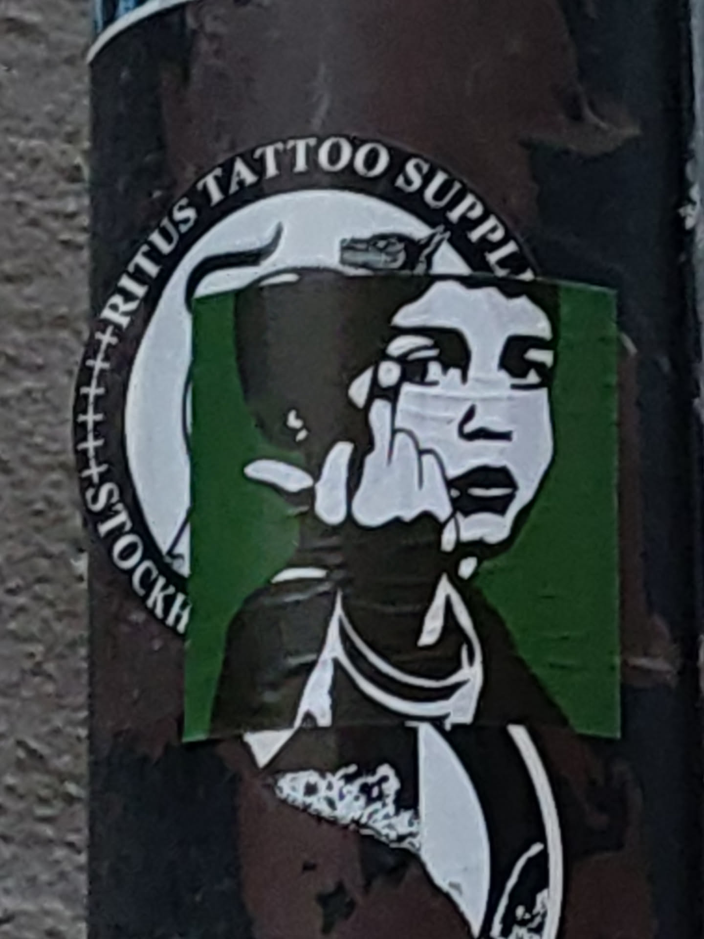 A sticker from Ritūs Tattoo Supply in Stockholm. The design depicts a person's face partially obscured by a dark object, with their middle finger raised. The background is green and the sticker has a circular border with the shop's name.