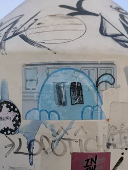 The sticker features a blue, ghost-like character with two dark eyes, appearing to be peeking out from behind a metro car window. Other graffiti and stickers surround it, adding to the urban aesthetic.