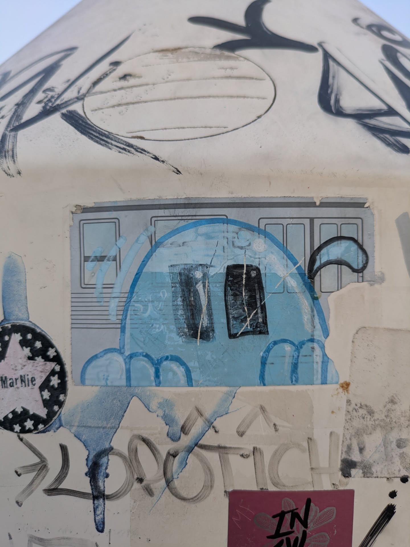 The sticker features a blue, ghost-like character with two dark eyes, appearing to be peeking out from behind a metro car window. Other graffiti and stickers surround it, adding to the urban aesthetic.