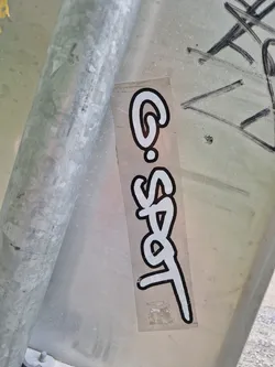 A vertical rectangular sticker featuring stylized white lettering on a clear background. The text appears to read 'G-Spot'.  The sticker is affixed to a metal pole, and there is some other graffiti visible on the surface near it. The style of the lettering is reminiscent of graffiti tags.