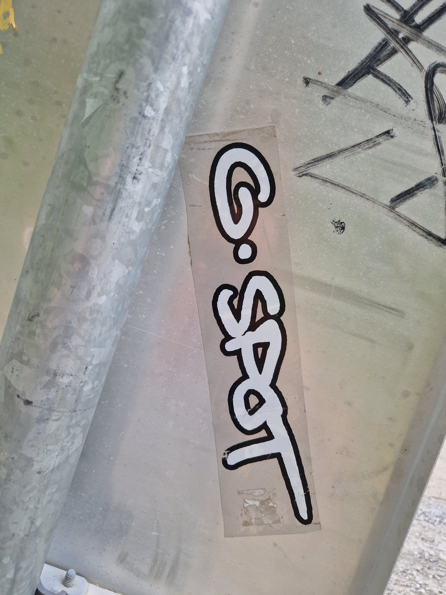 A vertical rectangular sticker featuring stylized white lettering on a clear background. The text appears to read 'G-Spot'.  The sticker is affixed to a metal pole, and there is some other graffiti visible on the surface near it. The style of the lettering is reminiscent of graffiti tags.