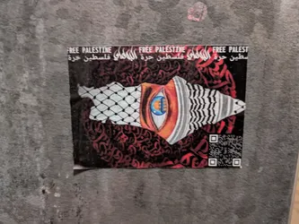 A sticker depicting a stylized eye and the map of Palestine, overlaid with Arabic calligraphy and a keffiyeh pattern. The text 'Free Palestine' is visible in both English and Arabic. A QR code is present in the bottom right corner.